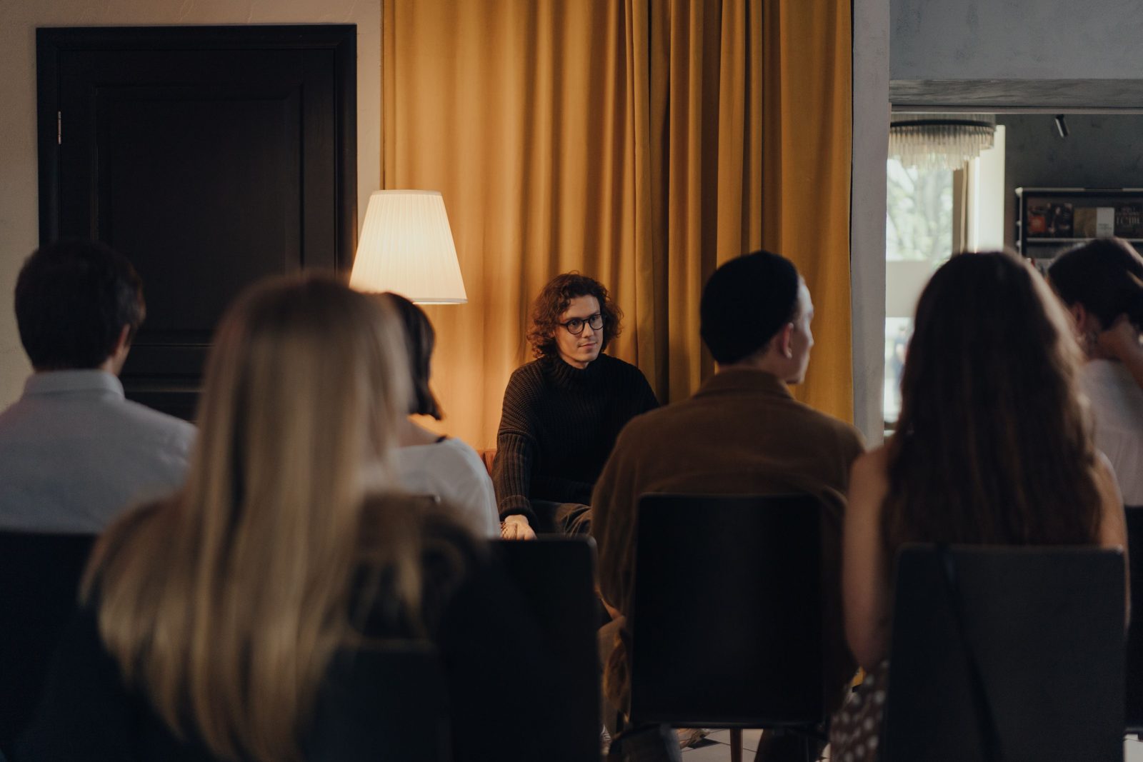 An audience seen from behind, listening to a speaker in a warmly lit room.