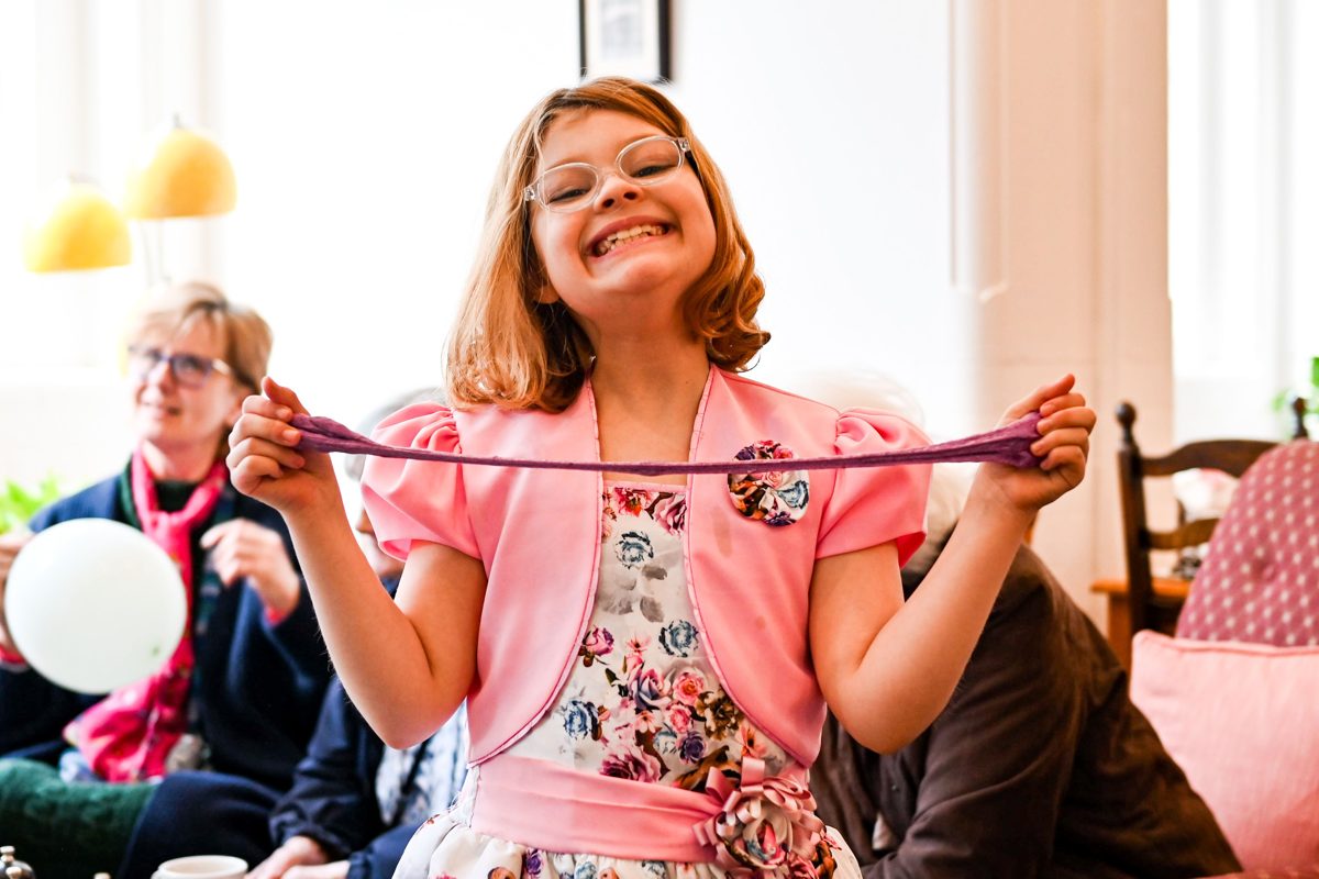 A smiling girl in a pink cardigan stretches a piece of slime between her hands at a party.
