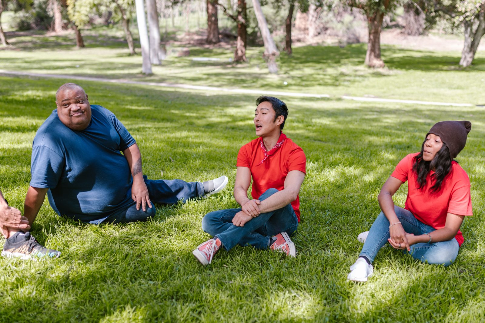 Three people sit together on the grass in a park, talking.