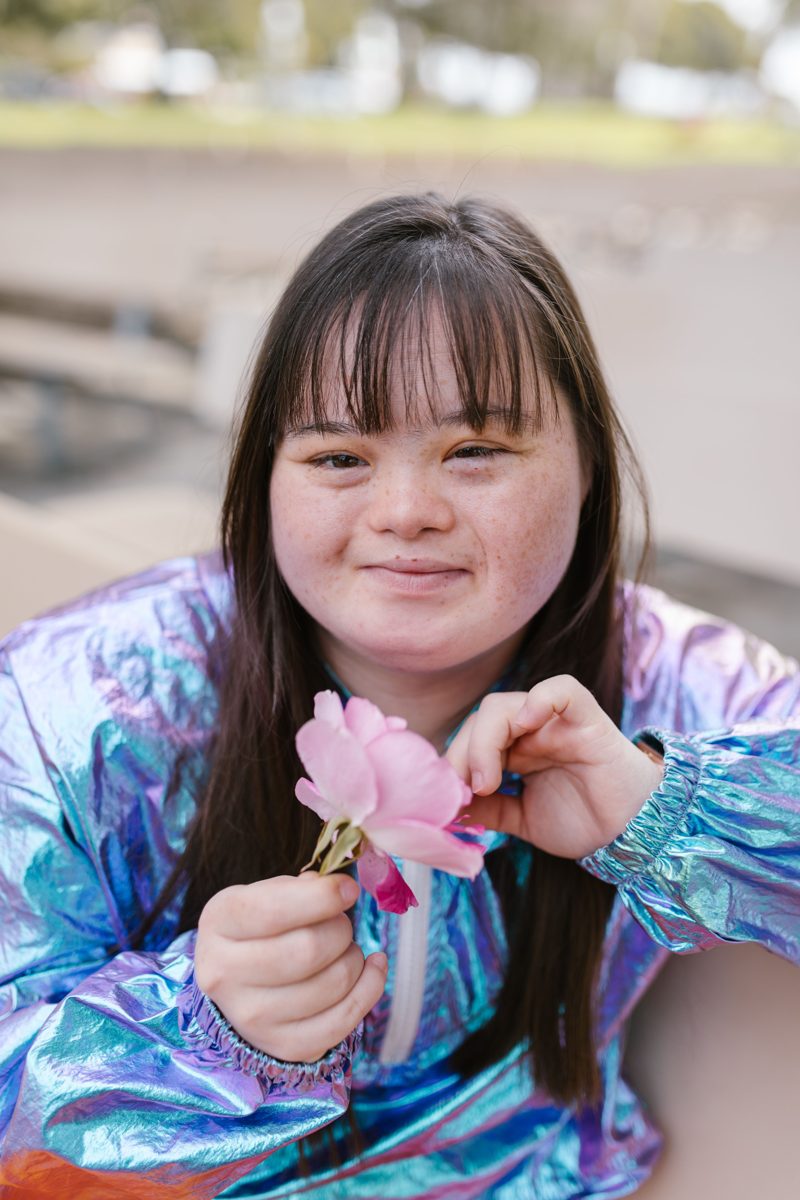 A young woman with Down syndrome smiles while holding a pink flower.
